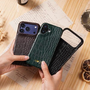 Op de afbeelding: Drie telefoonhoesjes met een krokodillenleertextuur worden getoond. De hoesjes zijn bruin, donkergroen en zwart. De groene hoes heeft gouden initialen. De hoesjes worden boven een houten oppervlak met een kaars en gedroogd fruit gehouden.