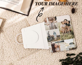 May include: A white phone case with a photo collage of family pictures. The case is open, revealing the photos. The background is a soft, cream-colored, textured surface. A book and a small potted plant are also visible.
