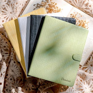 May include: A collection of leather-textured tablet cases in various colors, including yellow, white, gray, black, and light green. The light green case has a clasp closure and the name "Dominic" printed on it. The cases are arranged on a lace surface.