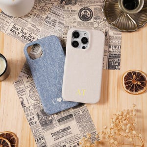 May include: Two phone cases, one light blue and one beige, with embroidered initials. The cases rest on a wooden surface with vintage newspaper clippings. Dried flowers, a candle, and a brass candle holder complete the scene.