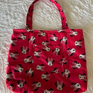 May include: Red tote bag featuring a pattern of Minnie Mouse faces with red and white polka dot bows. The bag has a matching red handle and a small pocket on the front. The bag is made of fabric and is designed for carrying items.