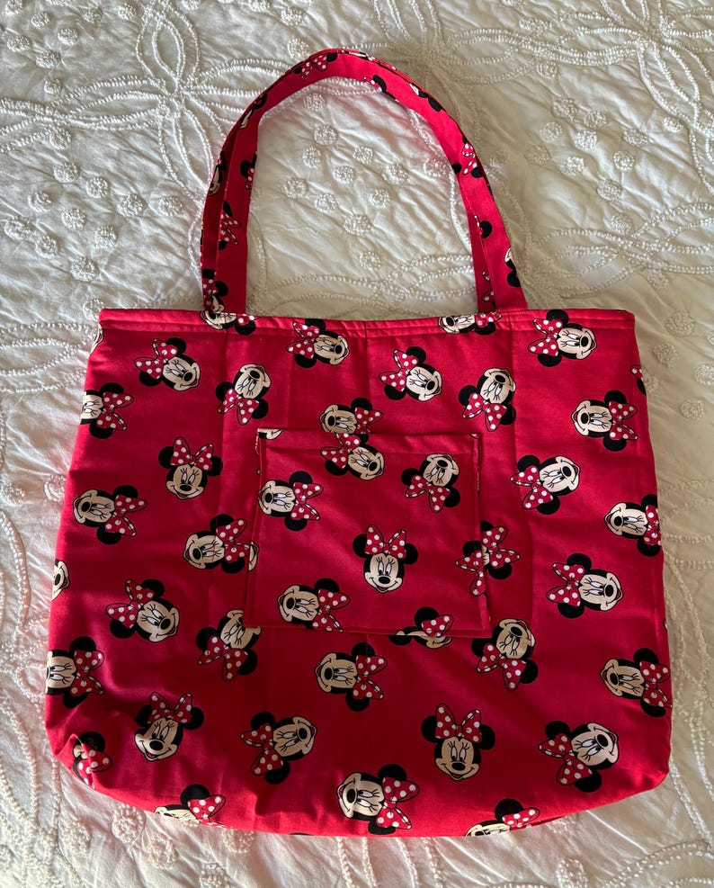 May include: Red tote bag featuring a pattern of Minnie Mouse faces with red and white polka dot bows. The bag has a matching red handle and a small pocket on the front. The bag is made of fabric and is designed for carrying items.