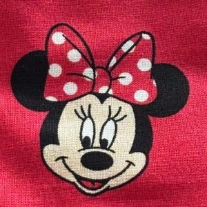 May include: Close-up of a red fabric featuring a Minnie Mouse design. The cartoon character has black ears, a smiling face, and a red bow with white polka dots. The background is a solid red color.