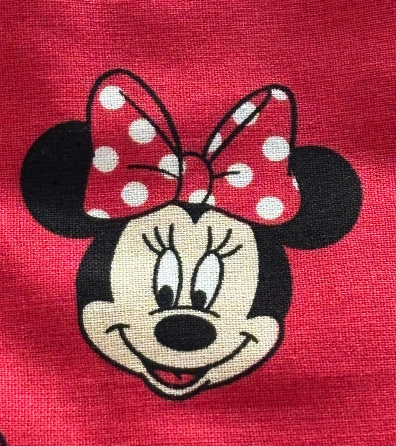 May include: Close-up of a red fabric featuring a Minnie Mouse design. The cartoon character has black ears, a smiling face, and a red bow with white polka dots. The background is a solid red color.