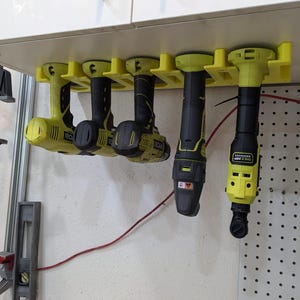 May include: A wall-mounted storage rack with five drill drivers hanging from it. The rack is made of white plastic and has a yellow accent. The drill drivers are all black and yellow and have different brands.