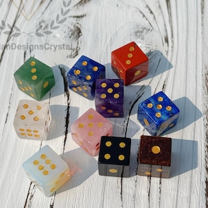 May include: A collection of gemstone dice in various colors, including green, red, blue, purple, pink, and black. Each die features gold-colored dots. The dice are arranged on a white wooden surface.