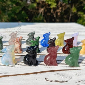 Puede incluir: Una colección de 15 pequeñas figuritas de lobo talladas en diferentes tipos de piedras pulidas. Las figuritas están todas en posición sentada con la cabeza levantada, y están dispuestas en semicírculo sobre una superficie de madera blanca.