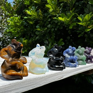 May include: A row of seven small carved elephant figurines made of different types of crystals. The elephants are all facing the same direction and have their trunks raised. The crystals are tiger's eye, opalite, obsidian, sodalite, aventurine, amethyst, and rose quartz.