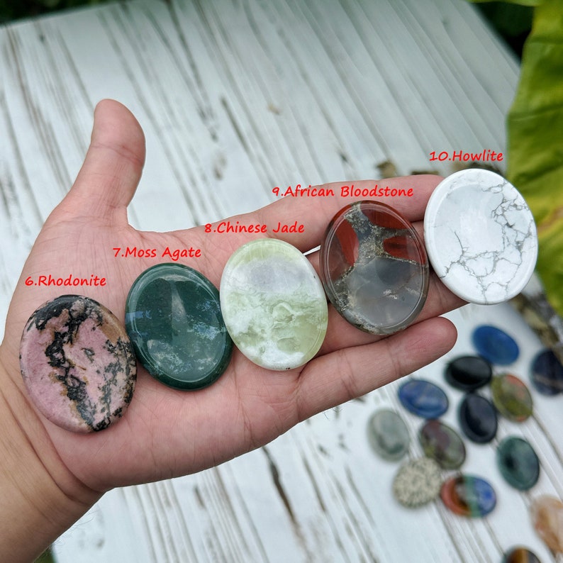 May include: Assortment of polished oval-shaped stones, including rhodonite, moss agate, Chinese jade, African bloodstone, and howlite. The stones are arranged in a row on a hand, with the names of each stone labeled in red text.