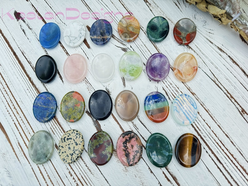 May include: Assortment of 18 oval-shaped polished stones, each with a unique color and pattern. The stones are arranged in rows on a white wooden surface.