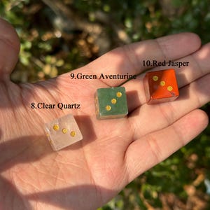 Puede incluir: Tres dados de piedras preciosas: cuarzo transparente, aventurina verde y jaspe rojo. Cada dado tiene puntos dorados. Los dados se sostienen en una mano sobre un fondo verde borroso. El texto de la imagen dice "8. Clear Quartz", "9. Green Aventurine" y "10. Red Jasper".
