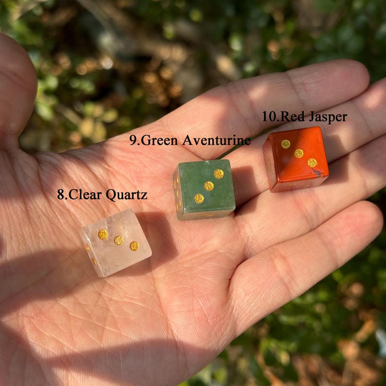 Puede incluir: Tres dados de piedras preciosas: cuarzo transparente, aventurina verde y jaspe rojo. Cada dado tiene puntos dorados. Los dados se sostienen en una mano sobre un fondo verde borroso. El texto de la imagen dice "8. Clear Quartz", "9. Green Aventurine" y "10. Red Jasper".