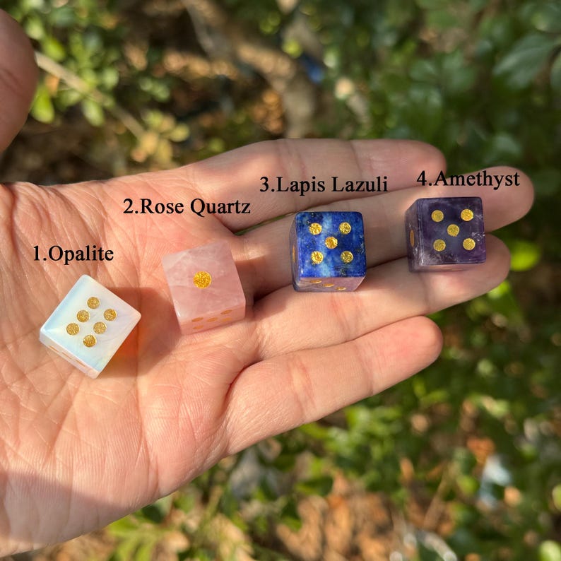 Puede incluir: Cuatro dados de piedras preciosas: Opalita, Cuarzo Rosa, Lapisl&aacute;zuli y Amatista. Cada dado es un cubo con puntos dorados que representan n&uacute;meros. Los dados se sostienen en una mano sobre un fondo verde borroso.