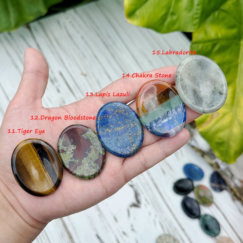 May include: A hand holding a set of seven oval-shaped polished stones. The stones are in various colors, including brown, green, blue, red, and gray. The stones are labeled with their names, including Tiger Eye, Dragon Bloodstone, Lapis Lazuli, Chakra Stone, and Labradorite.