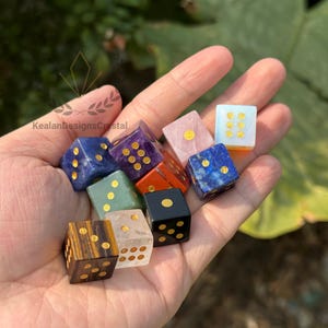 Puede incluir: Un conjunto de dados de piedras preciosas en varios colores, incluyendo azul, morado, verde y negro, con puntos dorados. Los dados se sostienen en una mano, mostrando sus superficies pulidas y patrones minerales &uacute;nicos. Un regalo &uacute;nico para los entusiastas de los juegos de mesa.