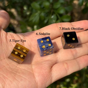 Puede incluir: Tres dados de piedras preciosas: Ojo de Tigre, Sodalita y Obsidiana Negra. Cada dado tiene puntos dorados. El dado de Ojo de Tigre es marr&oacute;n con rayas doradas. El dado de Sodalita es azul con puntos dorados. El dado de Obsidiana Negra es negro con puntos dorados.