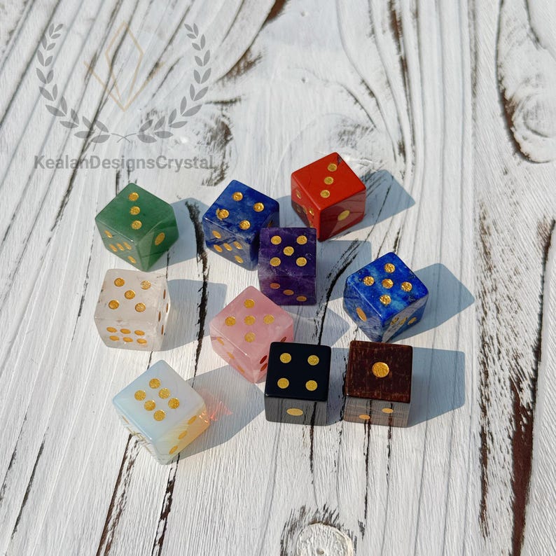 Puede incluir: Una colecci&oacute;n de dados de piedras preciosas en varios colores, incluyendo verde, rojo, azul, morado, rosa y negro. Cada dado presenta puntos dorados. Los dados est&aacute;n dispuestos sobre una superficie de madera blanca.