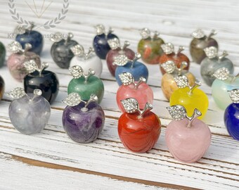 May include: A collection of gemstone apple figurines in various colors, including red, pink, green, and purple. Each apple has a silver-colored stem and leaf detail. The apples are arranged on a white wooden surface.