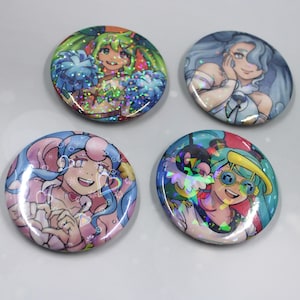 May include: Four round, colorful buttons featuring anime-style illustrations. The buttons showcase characters with vibrant hair and clothing, with a holographic, glittery effect. Each button has a unique design and color scheme, including pink, blue, green, and teal.