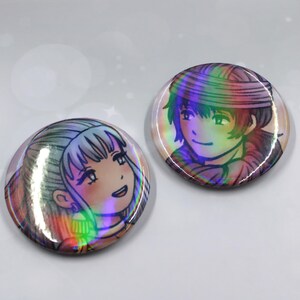 May include: Two round, holographic buttons featuring anime-style illustrations of characters with smiling faces. The buttons have a rainbow sheen, with the illustrations in black and shades of pink, blue, and green.