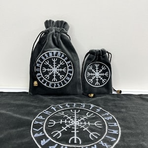 May include: Two dark grey velvet drawstring pouches and a matching fabric mat, each featuring an embroidered white Vegvisir symbol surrounded by a circular border of runic characters. The pouches have black drawstrings with small metallic beads.