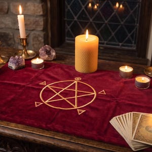 May include: A rectangular deep red velvet altar cloth featuring a gold embroidered pentagram symbol in the centre. The cloth is surrounded by several lit white and yellow candles in brass holders, scattered amethyst crystals, and tarot cards.