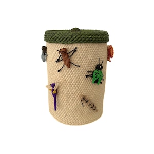 May include: A cylindrical beige woven container with a green rim and lid. The container is decorated with embroidered insects in various colors, including brown, green, and purple. The container is a decorative storage item.