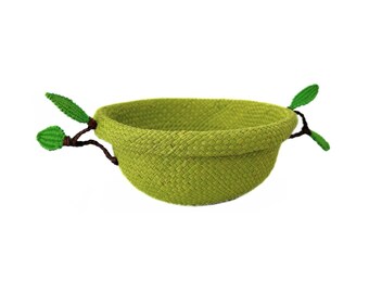 Puede incluir: Un cuenco tejido de color verde lima con forma redondeada. Dos elementos decorativos en forma de ramita con detalles de hojas verdes se extienden desde los lados. El cuenco está sobre un fondo blanco.