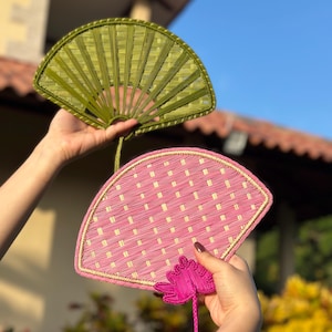 May include: Two hand fans, one olive green and one pink, are displayed against a bright blue sky. The fans are made of woven material, with a decorative floral accent on the pink fan. The fans are held up, showcasing their intricate design and vibrant colors.