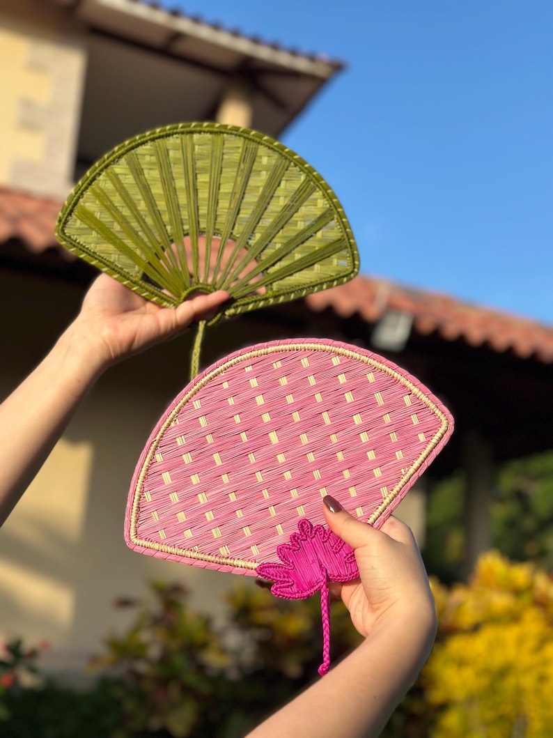 May include: Two hand fans, one olive green and one pink, are displayed against a bright blue sky. The fans are made of woven material, with a decorative floral accent on the pink fan. The fans are held up, showcasing their intricate design and vibrant colors.