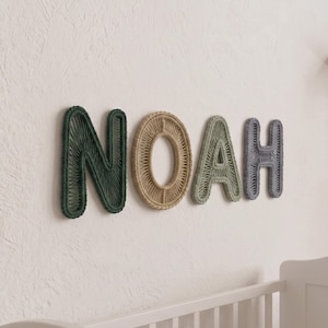 Może przedstawiać: Zestaw czterech liter ściennych tworzących imię NOAH. Każda litera wykonana jest z drucianego stelaża owiniętego kolorowym sznurkiem. Litera N jest ciemnozielona, O jest beżowe, A jest szałwiowozielone, a H ma kolor przygaszonego błękitu.