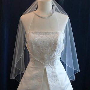 Könnte beinhalten: Ein weißes Brautkleid mit besticktem Oberteil und fließendem Rock. Ein durchsichtiger Schleier ist über das Kleid drapiert, und eine glitzernde Halskette ziert den Hals der Schaufensterpuppe. Das Kleid wird auf einer Schaufensterpuppe präsentiert.