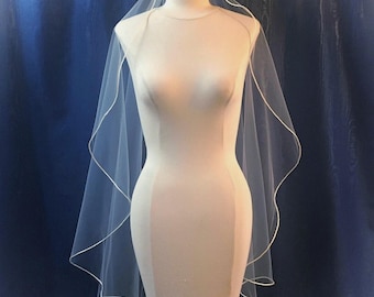 May include: A sheer white bridal veil draped over a mannequin torso. The veil features a delicate, thin white satin ribbon trim along the entire scalloped edge, creating a soft, flowing silhouette that cascades down the sides of the form.