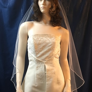 K&ouml;nnte beinhalten: Elfenbeinfarbenes Brautkleid ohne Tr&auml;ger mit floralen Stickereien und passendem Schleier. Das Kleid hat ein eng anliegendes Oberteil und einen flie&szlig;enden Rock. Die Brautrobe wird auf einer Schaufensterpuppe vor dunklem Hintergrund pr&auml;sentiert.