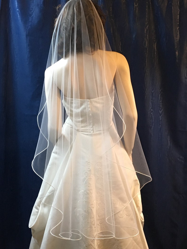 K&ouml;nnte beinhalten: Ein wei&szlig;es Brautkleid mit herzf&ouml;rmigem Ausschnitt und flie&szlig;endem Rock. Ein durchsichtiger, wei&szlig;er Schleier mit zarter Bord&uuml;re f&auml;llt &uuml;ber den R&uuml;cken. Das Kleid hat dezente gestickte Details.