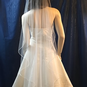 K&ouml;nnte beinhalten: Ein wei&szlig;es Brautkleid mit flie&szlig;endem Rock und einem durchsichtigen, mehrschichtigen Schleier. Der Schleier hat eine zarte, wellenf&ouml;rmige Bord&uuml;re. Das Kleid hat ein figurbetontes Oberteil und einen Knopfverschluss am R&uuml;cken. Der Hintergrund ist ein dunkelblauer Vorhang.