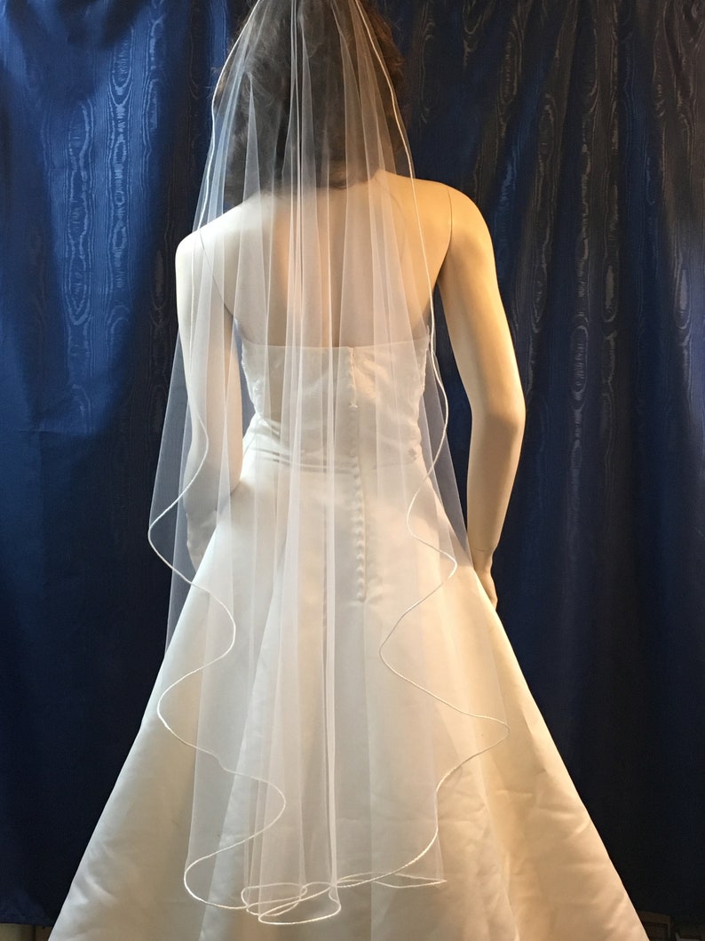K&ouml;nnte beinhalten: Ein wei&szlig;es Brautkleid mit flie&szlig;endem Rock und einem durchsichtigen, mehrschichtigen Schleier. Der Schleier hat eine zarte, wellenf&ouml;rmige Bord&uuml;re. Das Kleid hat ein figurbetontes Oberteil und einen Knopfverschluss am R&uuml;cken. Der Hintergrund ist ein dunkelblauer Vorhang.