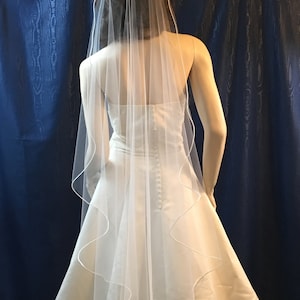 K&ouml;nnte beinhalten: Ein wei&szlig;er Brautschleier mit gewelltem Rand und schlichtem Design. Der Schleier ist &uuml;ber einem wei&szlig;en Brautkleid drapiert und erzeugt einen klassischen und romantischen Look. Der Schleier besteht aus einem transparenten, leichten Stoff.