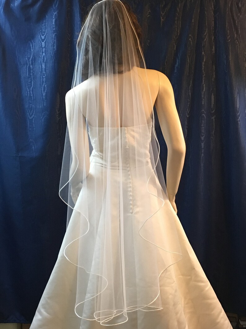 K&ouml;nnte beinhalten: Ein wei&szlig;er Brautschleier mit gewelltem Rand und schlichtem Design. Der Schleier ist &uuml;ber einem wei&szlig;en Brautkleid drapiert und erzeugt einen klassischen und romantischen Look. Der Schleier besteht aus einem transparenten, leichten Stoff.