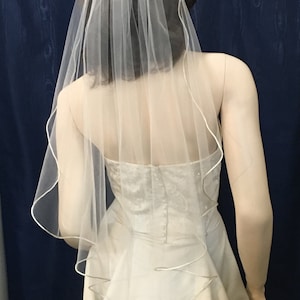 Könnte beinhalten: Ein weißer Braut Schleier mit Satin-Rand. Der Schleier ist einlagig und hat ein schlichtes Design.