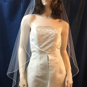 K&ouml;nnte beinhalten: Elfenbeinfarbenes tr&auml;gerloses Brautkleid mit besticktem Oberteil und flie&szlig;endem Rock. Ein durchsichtiger, wei&szlig;er Schleier mit zarter Bord&uuml;re ist &uuml;ber Kopf und Schultern der Schaufensterpuppe drapiert. Das Kleid hat einen Schlitz vorne.