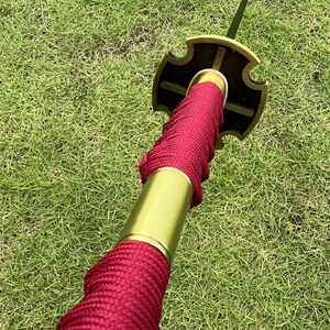May include: Close-up of a red and gold katana sword with a black and gold tsuba. The sword is wrapped in red cord and has a gold hilt.