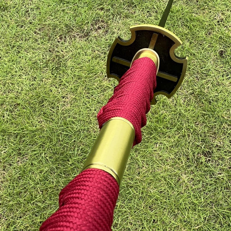 May include: Close-up of a red and gold katana sword with a black and gold tsuba. The sword is wrapped in red cord and has a gold hilt.