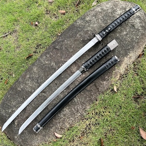 Op de afbeelding: Drie zwaarden in Japanse stijl met zilveren bladen en zwarte handvatten en scheden worden getoond op een grijze steen. De handvatten zijn gewikkeld in een zwart materiaal met textuur. De bladen zijn scherp en de algehele esthetiek is strak en traditioneel.
