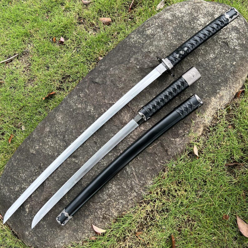 Op de afbeelding: Drie zwaarden in Japanse stijl met zilveren bladen en zwarte handvatten en scheden worden getoond op een grijze steen. De handvatten zijn gewikkeld in een zwart materiaal met textuur. De bladen zijn scherp en de algehele esthetiek is strak en traditioneel.