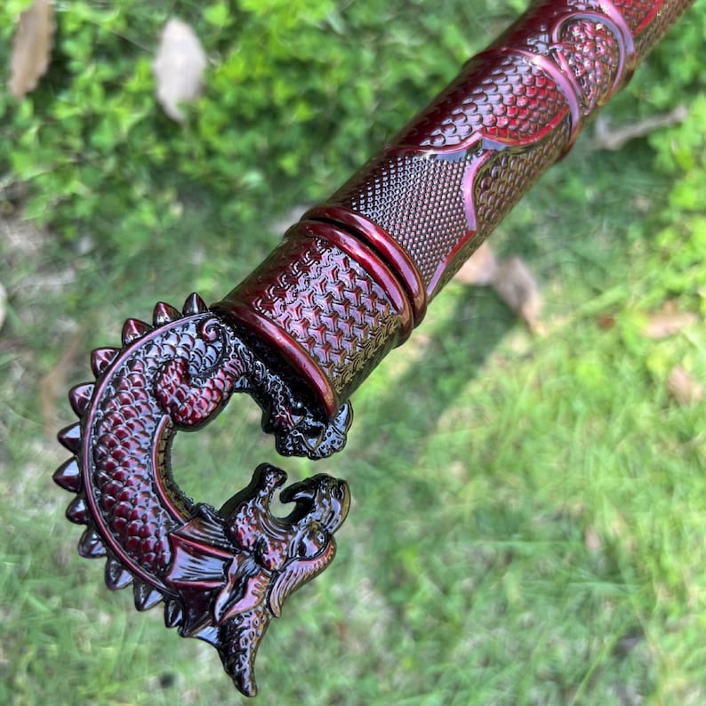 Peut inclure: Une poign&eacute;e de canne m&eacute;tallique rouge fonc&eacute; repr&eacute;sentant une t&ecirc;te de dragon sculpt&eacute;e avec des textures d'&eacute;cailles complexes. Le manche cylindrique attach&eacute; &agrave; la poign&eacute;e pr&eacute;sente un motif en losange r&eacute;p&eacute;titif et une finition brillante.