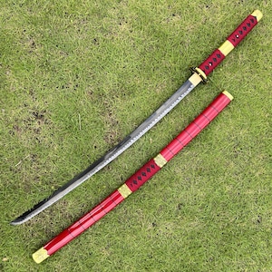 May include: A black and red katana sword with a gold-colored hilt and scabbard. The sword is lying on a green grassy surface.