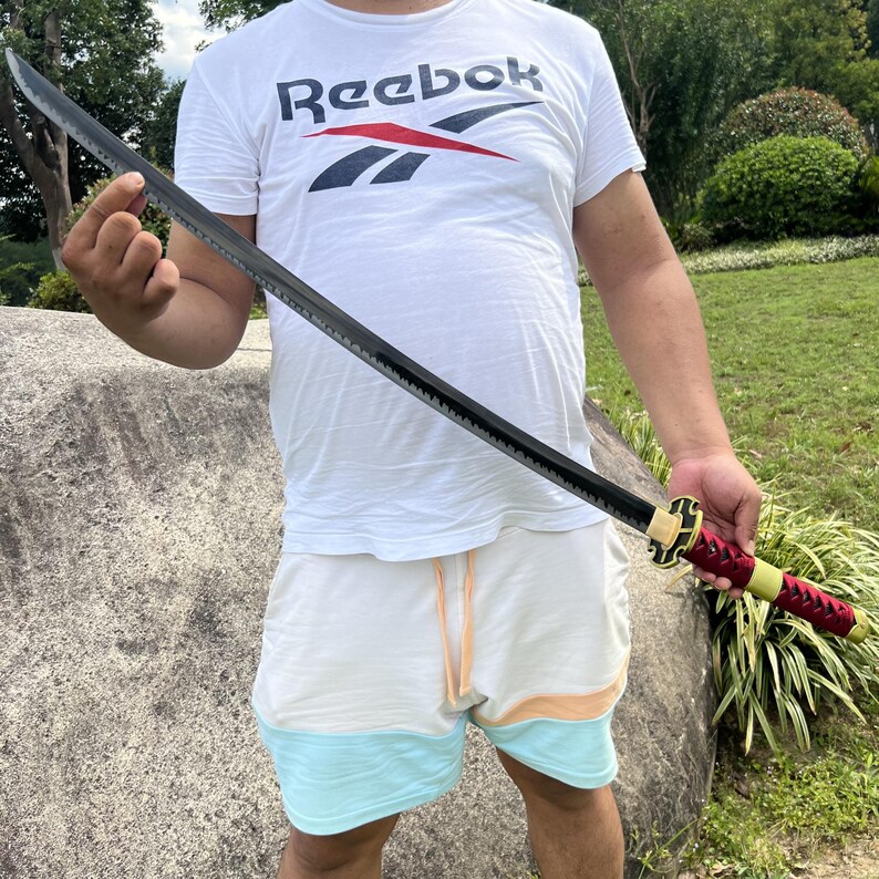 May include: A person holding a black katana sword with a red and gold handle. The sword is sharp and has a black scabbard.