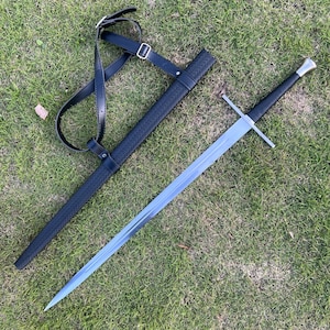 May include: A long, silver sword with a black handle and crossguard, resting on green grass. The sword is next to a black scabbard with a textured pattern and a black leather shoulder strap.