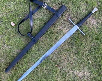 May include: A long, silver sword with a black handle and crossguard, resting on green grass. The sword is next to a black scabbard with a textured pattern and a black leather shoulder strap.